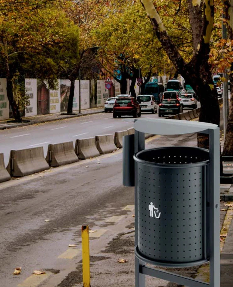 Papeleira Urbana Metálica para Exterior coberta com cinzeiro, instalada em ambiente urbano, em uma estrada com árvores.