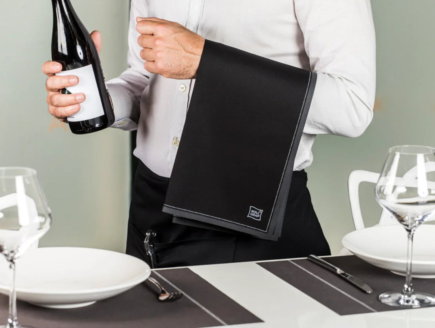 Rolo de Pano de Hotelaria Roll Drap em uso por garçom segurando garrafa e pano de mesa na preparação de mesa.
